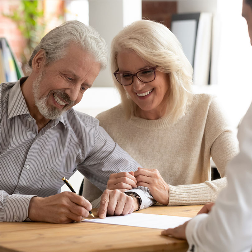 A smiling man signs a document as a smiling woman holds his arm.