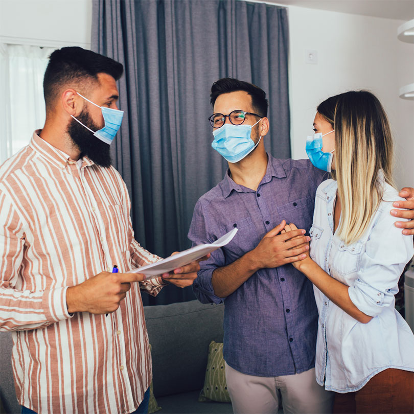 A couple wearing masks hold hands beside a man wearing a mask and holding a stack of papers.